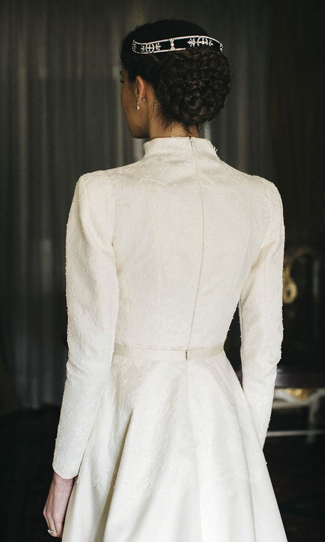 Carlota con vestido blanco y tiara, vista desde atrás en una habitación elegante.
