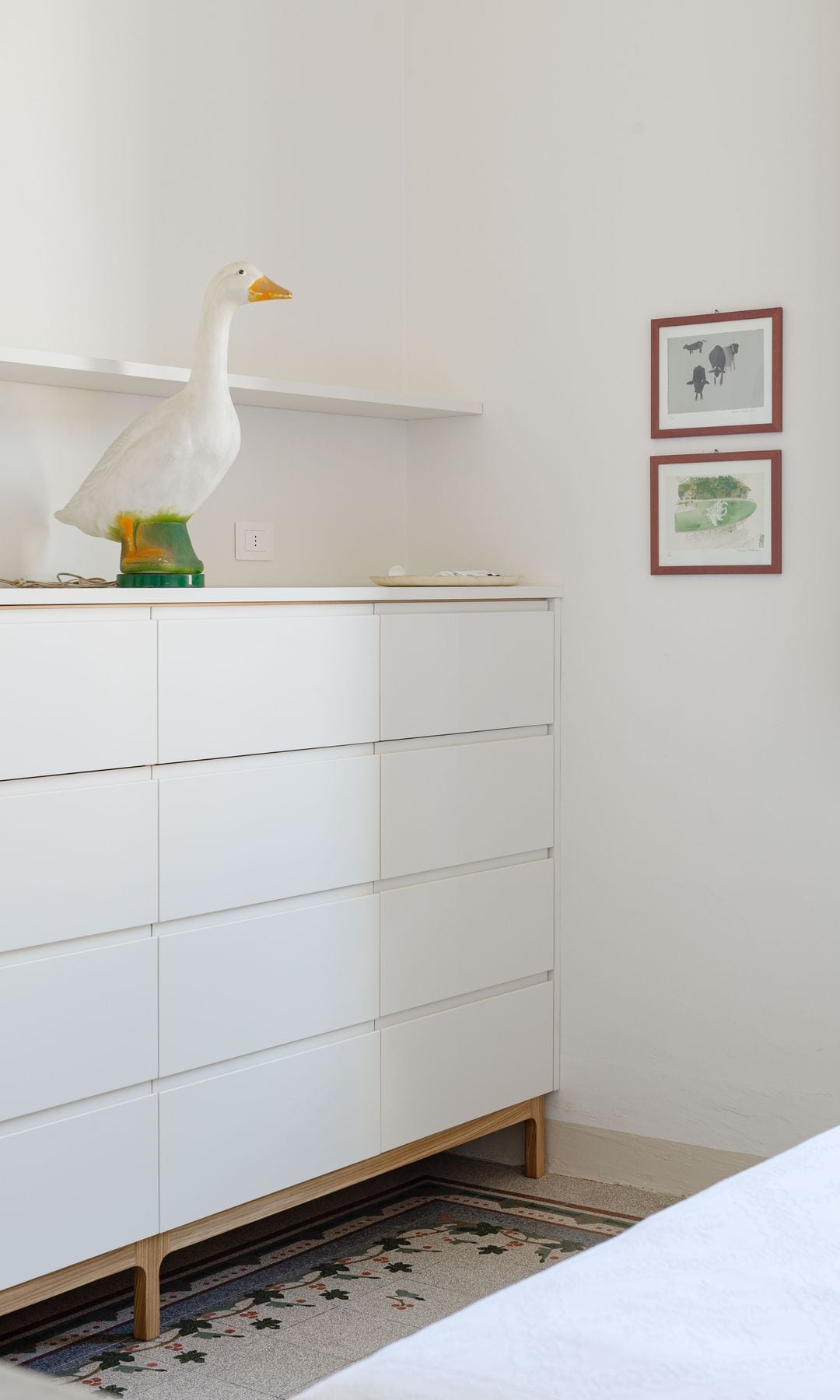 Cómoda del dormitorio con figura de una oca, cuadros en la pared, balda, suelo de terrazo 