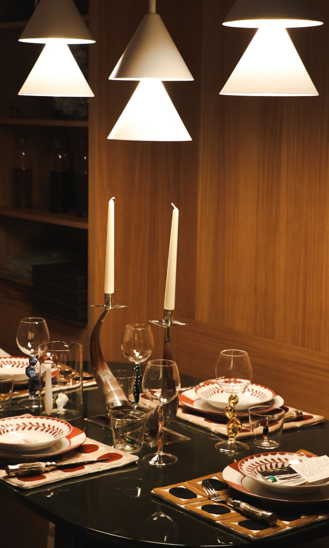 Mesa de color oscuro, montada con manteles individuales, copas, candelabros, vajilla blanca con motivos rojos, lámparas de techo