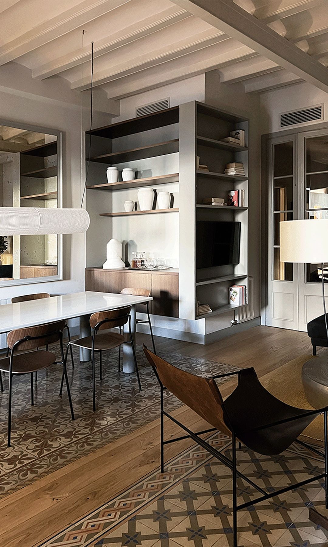 Salón comedor con vigas en el techo, suelo de baldosas hidráulicas, silla de cuero, lámpara de techo blanca, lámpara de pie, baldas en la pared, aparador