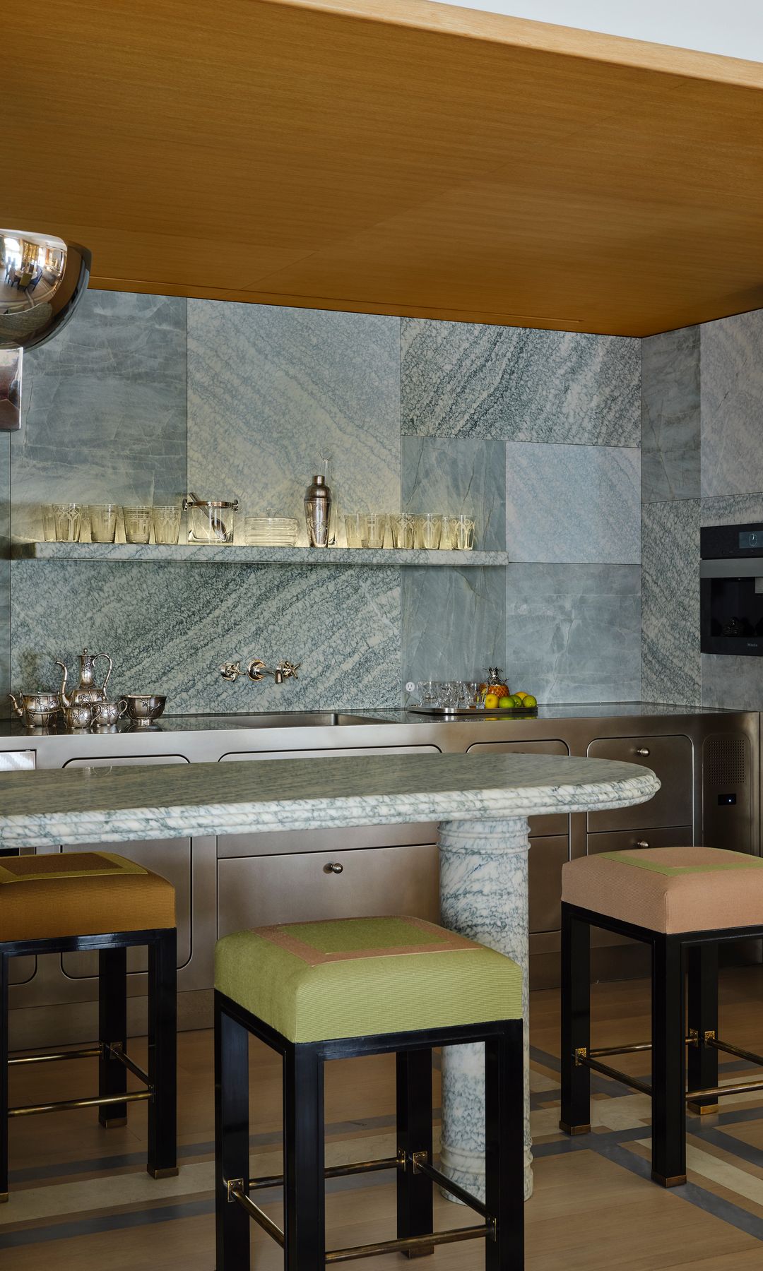 Cocina de acero, con isla central de piedra, paredes del mismo material, techo de madera, taburetes altos con asientos tapizados de distintos colores, 