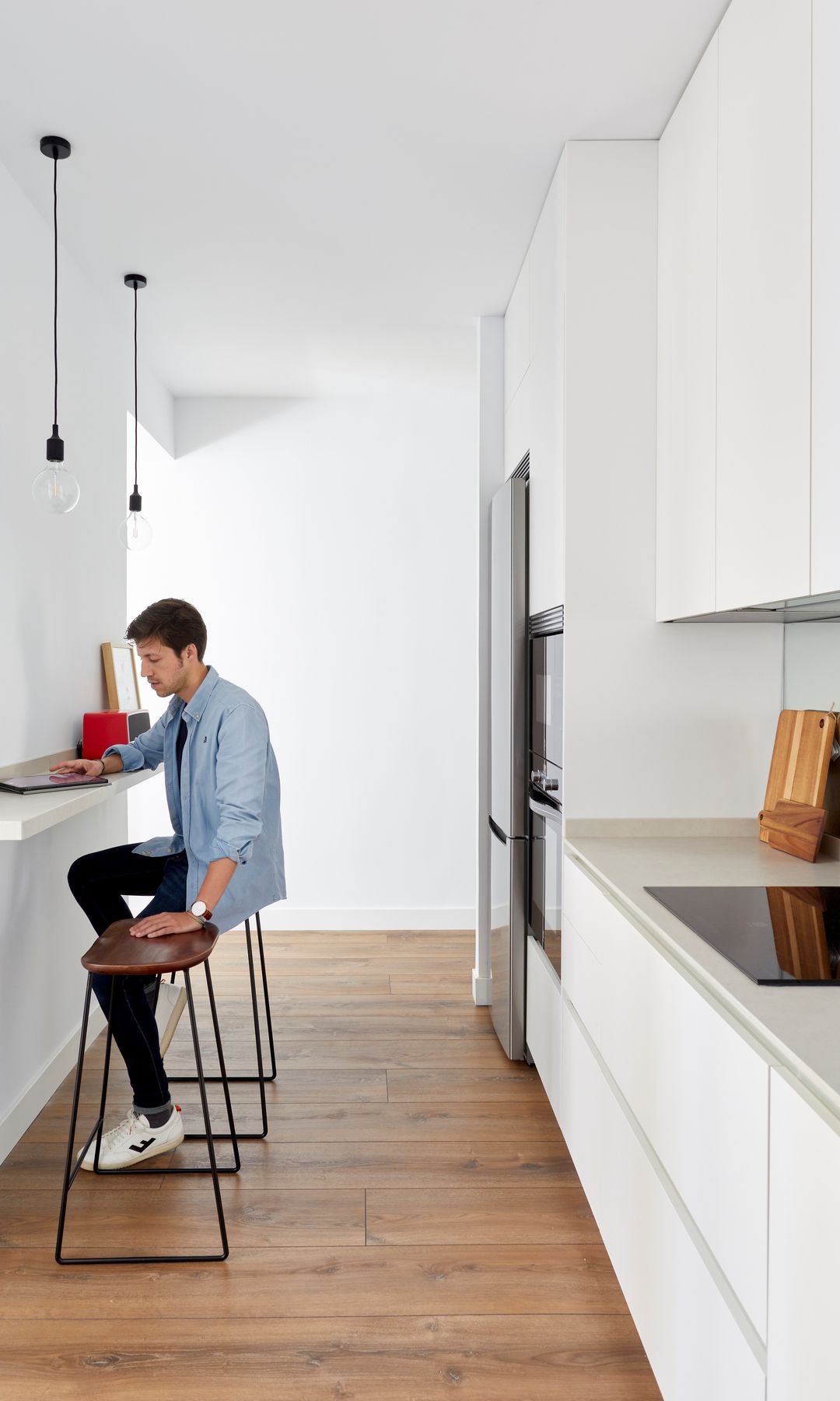 Cocina alargada con muebles blancos, encimera blanca, pared con mesa volada con taburetes, chico sentado, suelo de madera