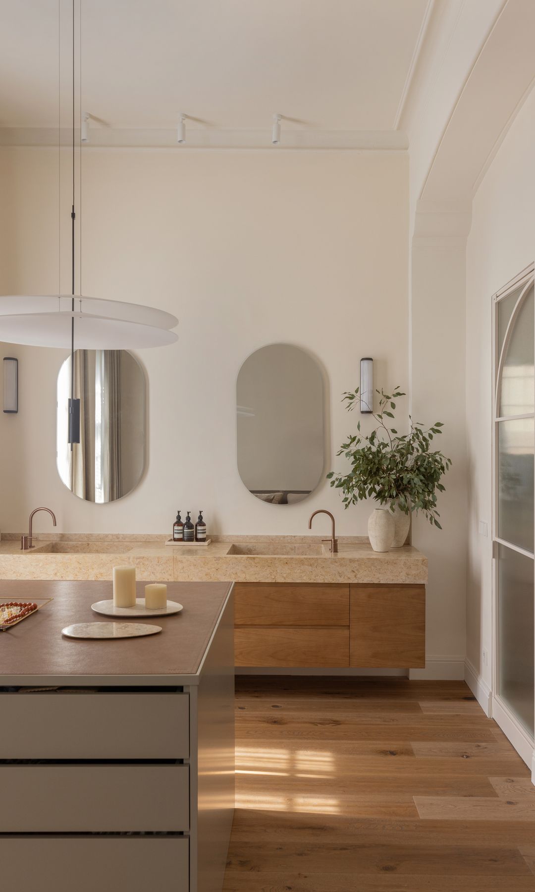 Baño XL con cómoda central, mueble bajolavabo doble de madera, espejos, jarrón con ramas verdes, apliques a ambos lados de los espejos, puerta de cristal para la ducha  
