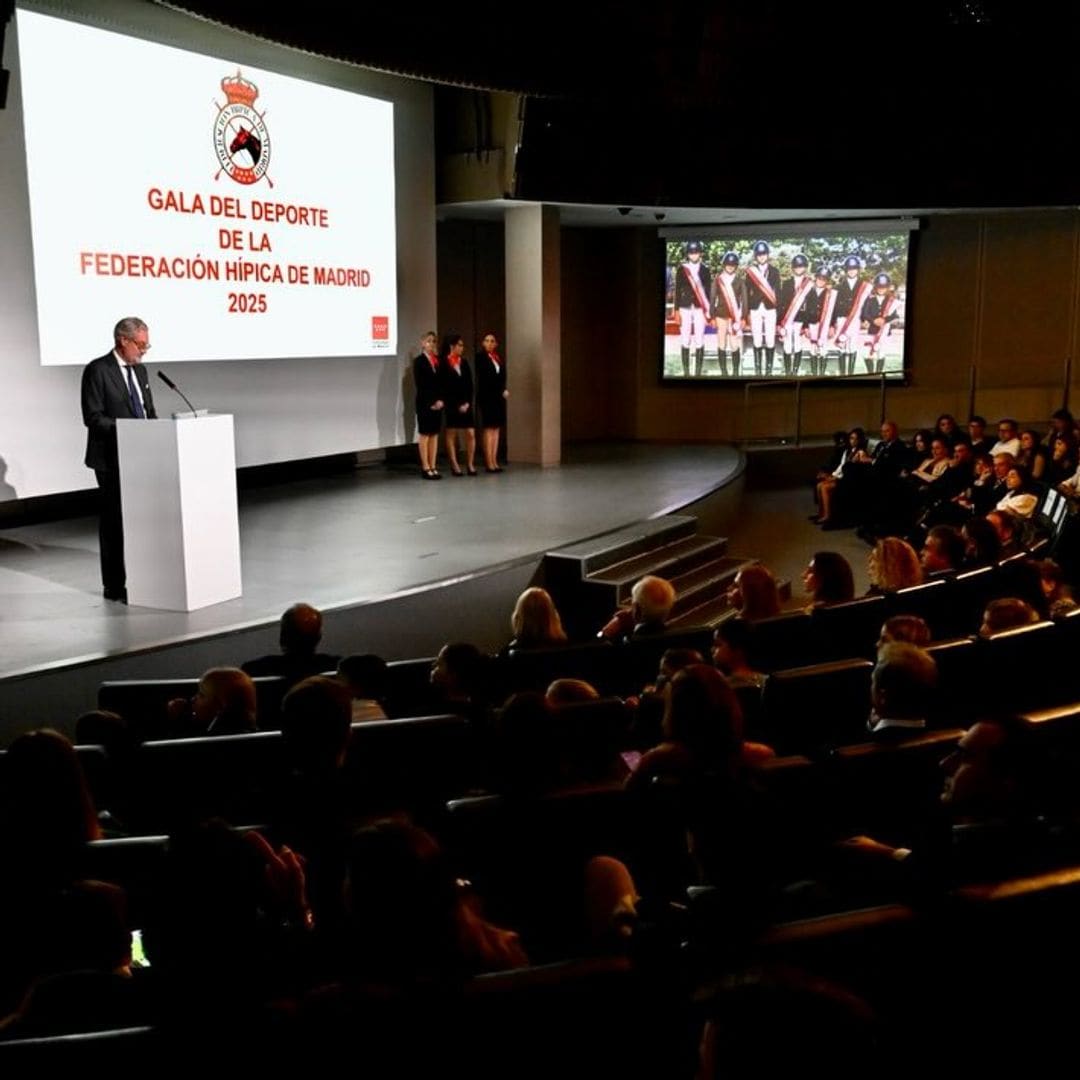Hombre en traje dando discurso en evento de gala, con dos mujeres en trajes negros al lado.