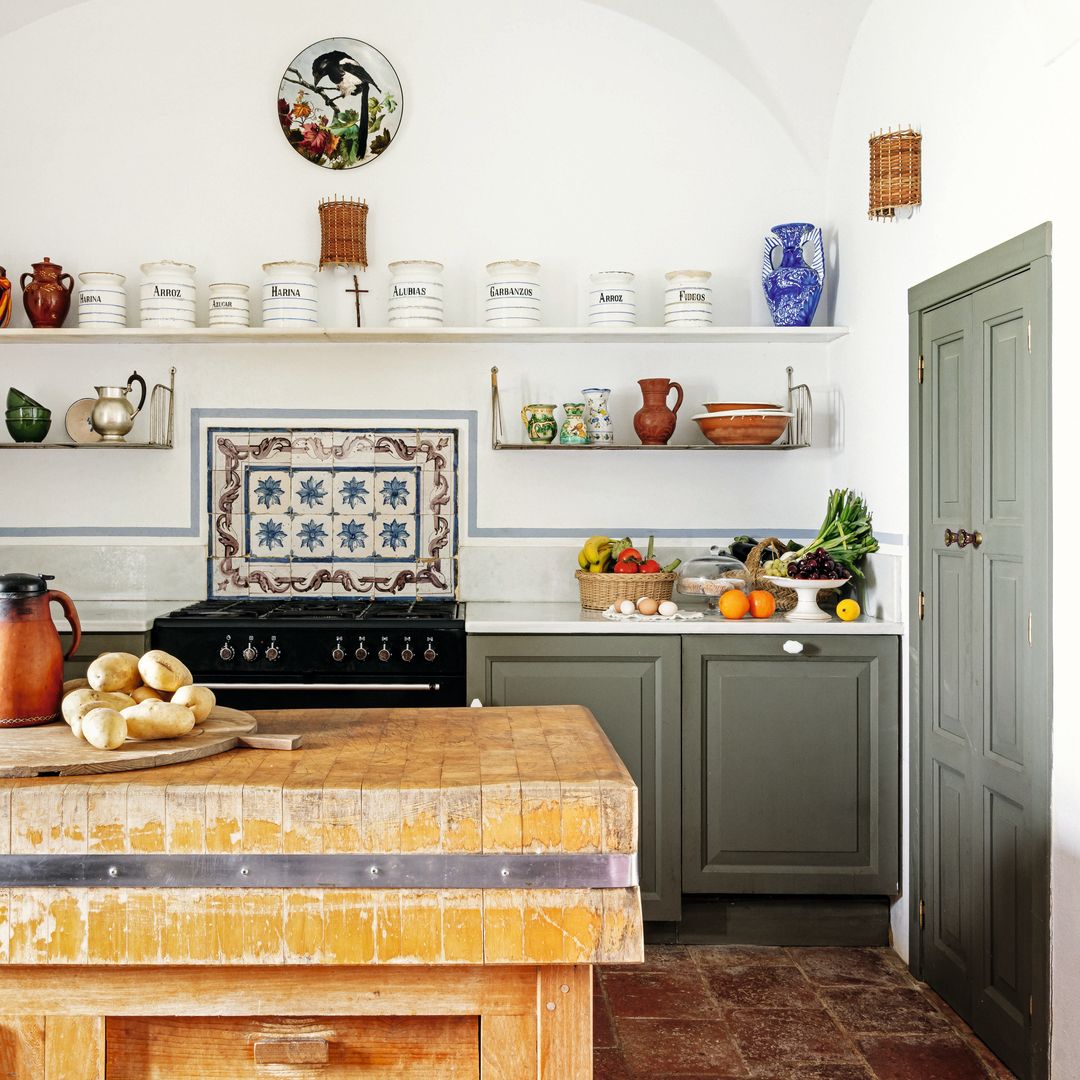 Cocina rústica con mesa de madera, estantes con jarras, y frutas en la encimera.