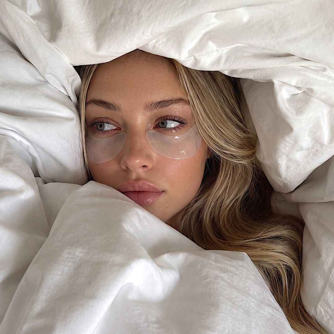 Primer plano de chica rubia con parches en los ojos rodeada por un edredón blanco