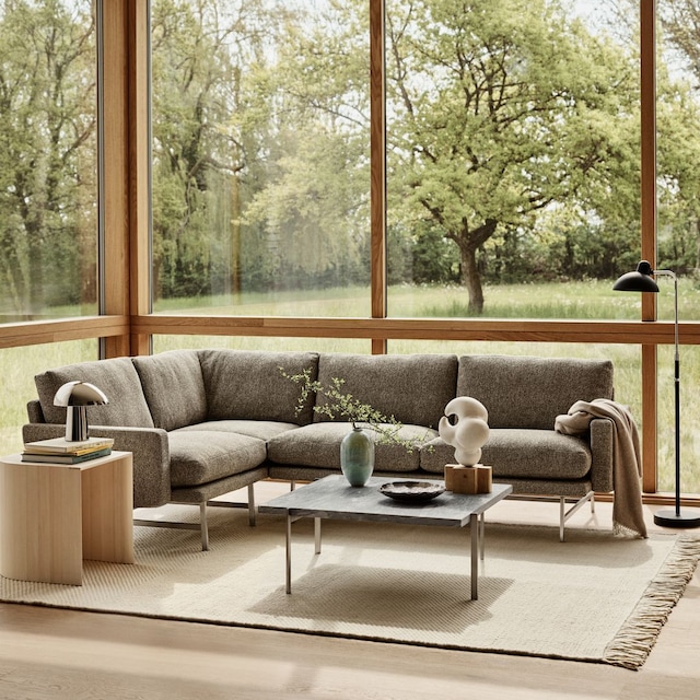 Salón moderno con sofá gris, alfombra de flecos, lámparas de pie y de sobremesa, cortinas blancas y grandes ventanales de madera