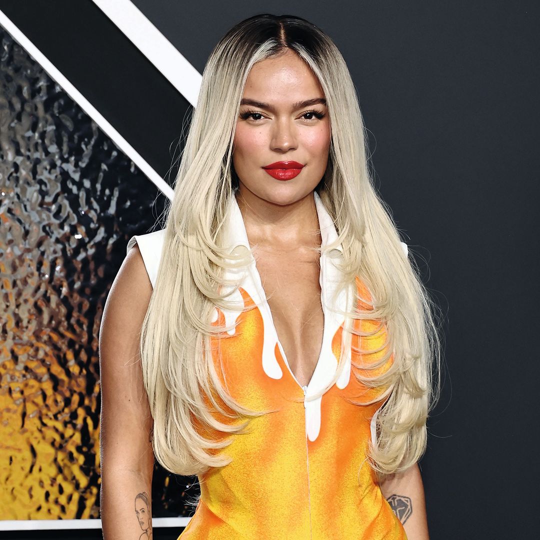 Karol G con pelo largo rubio, vestido naranja y amarillo, y labios rojos