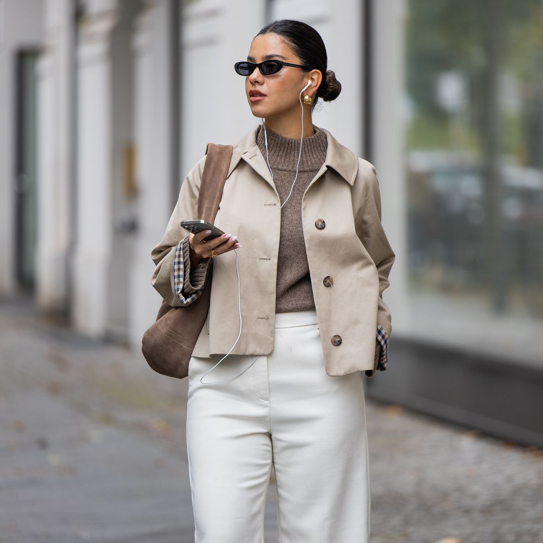 BERLÍN, ALEMANIA - 8 DE OCTUBRE: Bella Emar con un iPhone, auriculares con cable de Apple, chaqueta tipo trench de Sézane, jersey de punto y pantalones acampanados blancos, botas favorecedoras y bolso de ante marrón de Liebeskind el 8 de octubre de 2025 en Berlín, Alemania. (Foto de Christian Vierig/Getty Images)