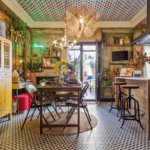 Cocina con office con suelo de baldosas geométricos, mesa de madera, alfombra de fibras vegetales, alacena amarilla, taburetes de hierro, puffs rojos y plantas