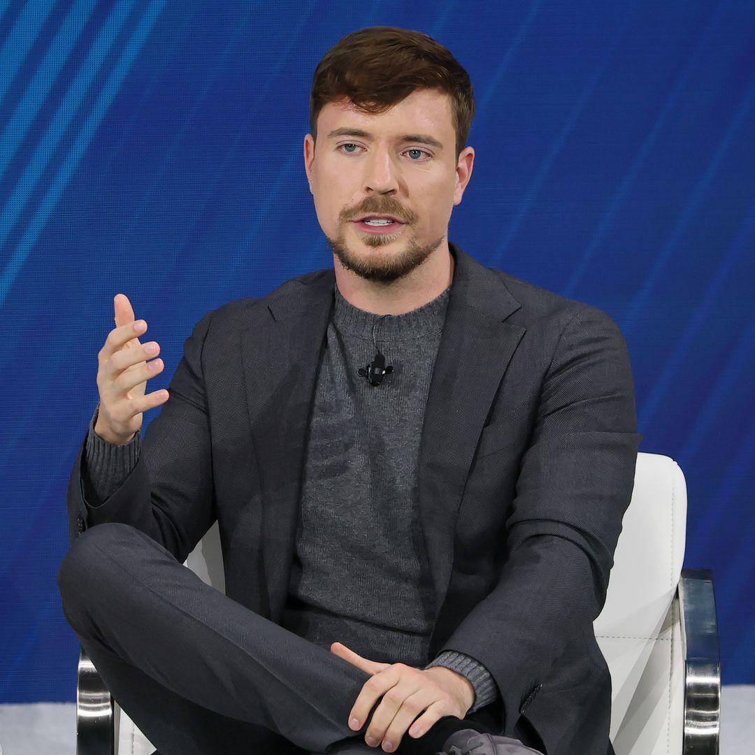 MrBeast en una silla, con traje gris y camiseta gris, habla en un evento.