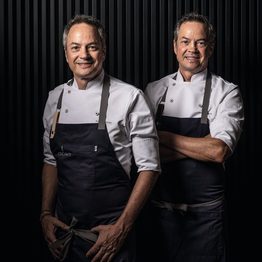 Los Hermanos Torres sonrientes posan juntos, uno con delantal que dice 'COCINA'.