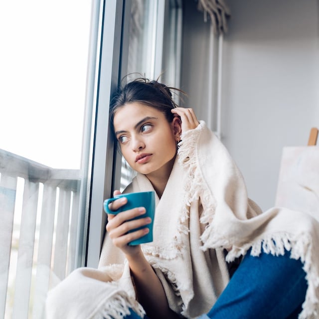 mujer tomando un té cubierta con una manta, mirando por la ventana