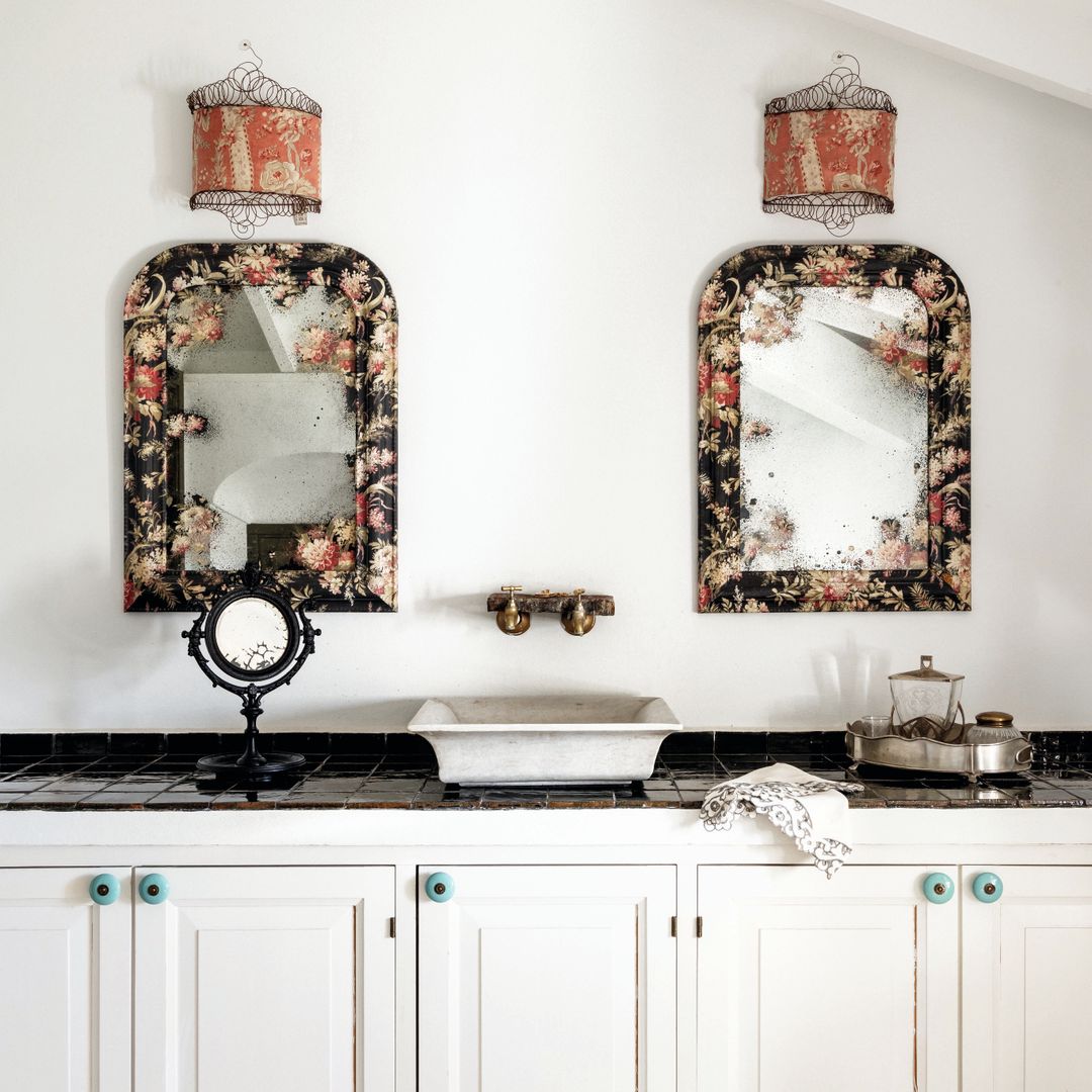 Baño elegante con espejos florales, lámparas vintage, y gabinetes blancos.