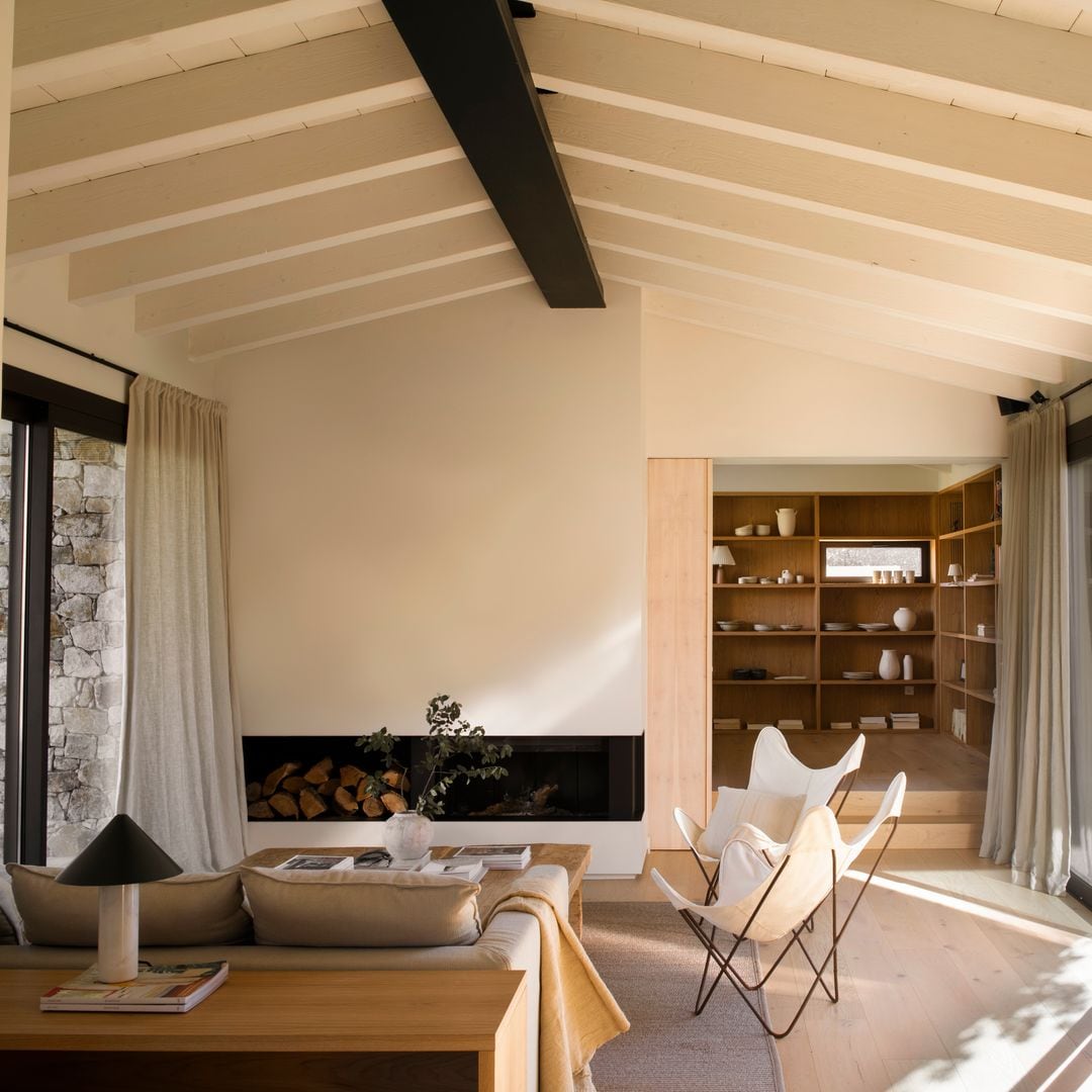 Salón con chimenea, abierto al rincón de lectura, vigas de madera, ventanales con salida al jardín, mesa de centro de madera, butacas de lona blancas, cortinas beiges, aparador de madera 