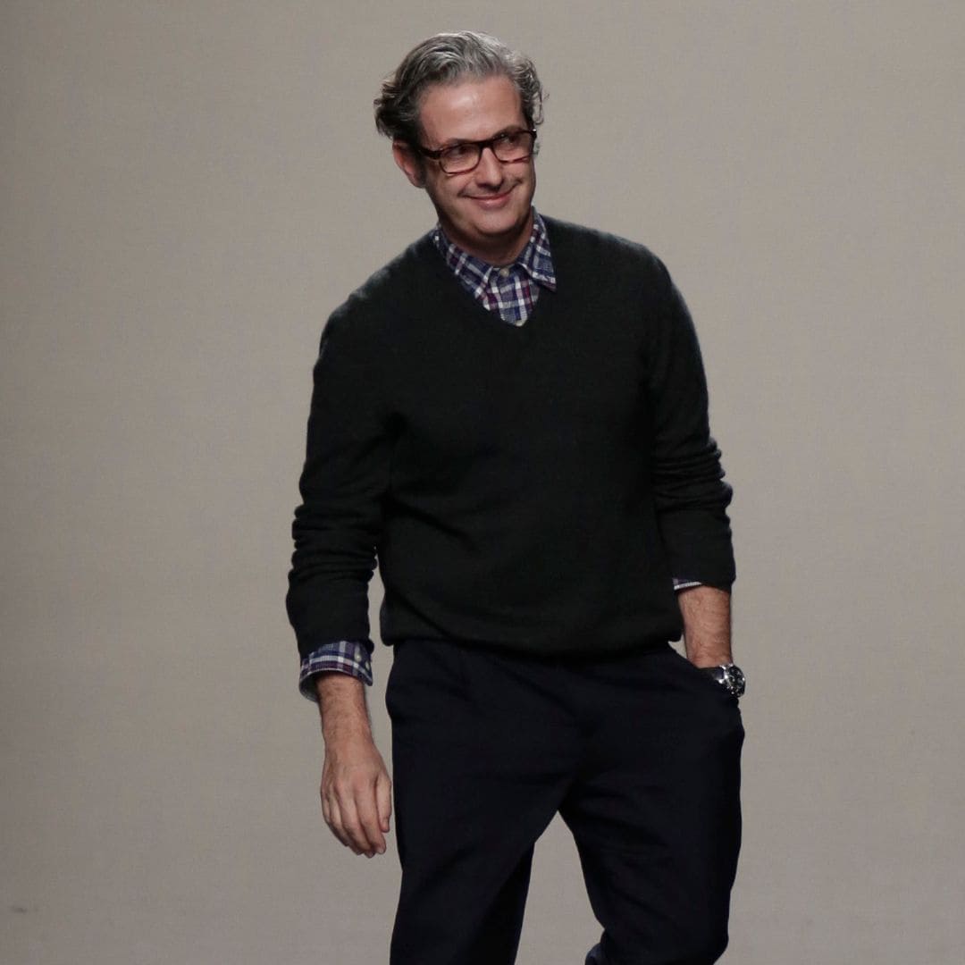 Miguel Palacio con cabello gris y gafas, vestido con suéter negro y pantalones, caminando en pasarela.