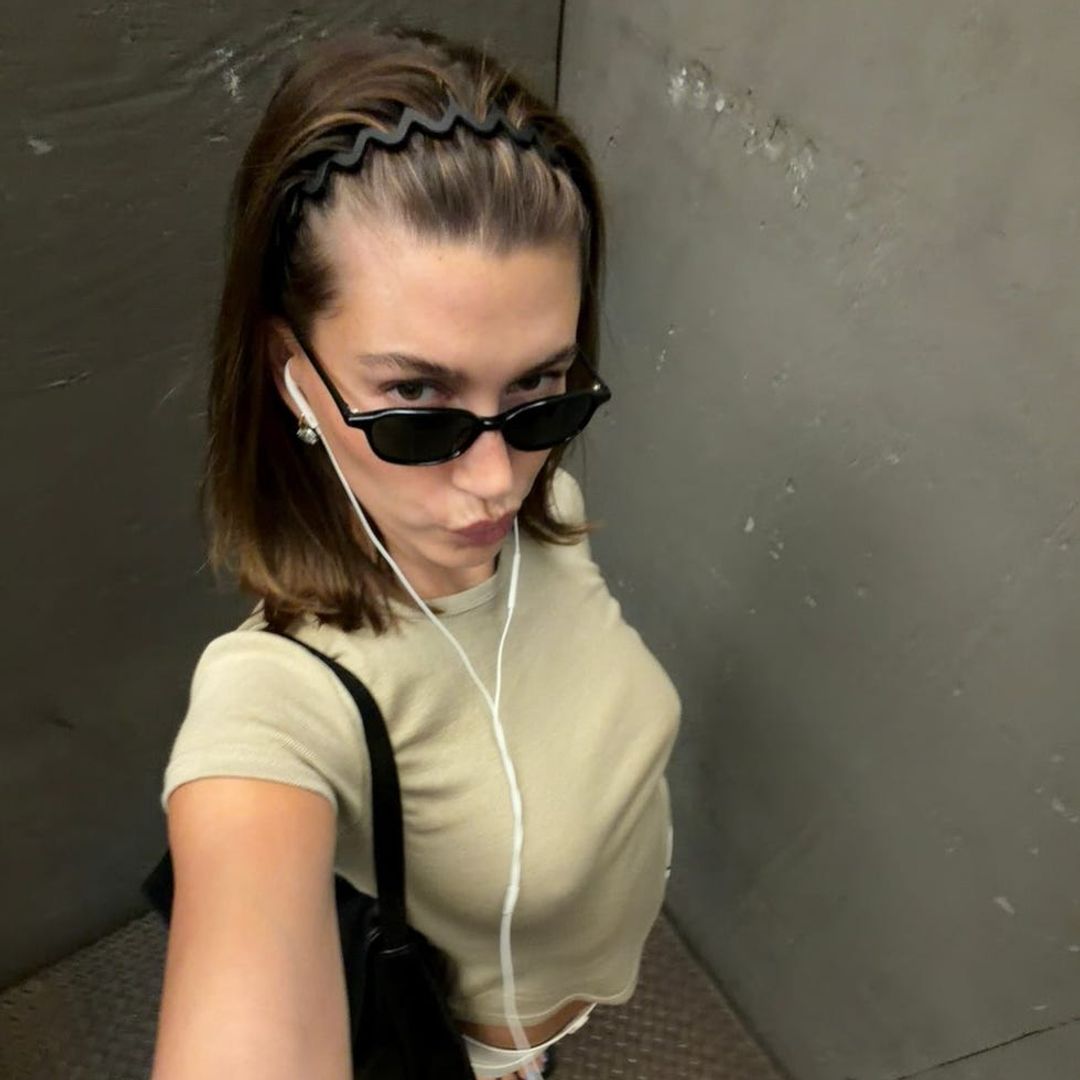 Hailey Bieber con gafas de sol, camiseta en tono crudo, bolso negro y auriculares