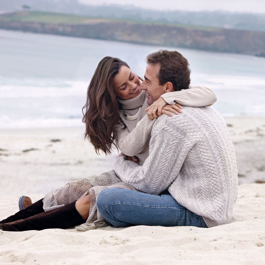 María Palacios con botas y Alessandro Lequio con jeans abrazados en la playa mirando al mar