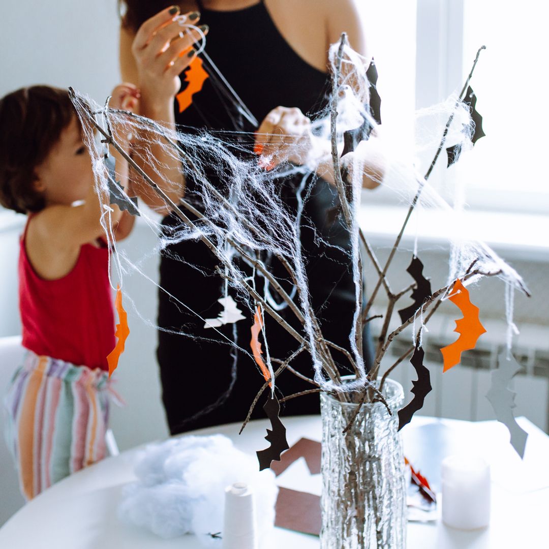Madre con hija realizando manualidades de Halloween con un jarrón, ramas secas, telarañas y pequeños murciélagos en negro y naranja, que contrastan con la casa en blanco.