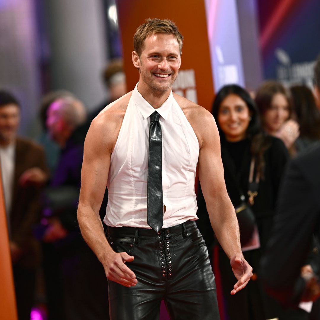 Alexander Skarsgård en traje de cuero negro y camisa blanca con corbata negra.