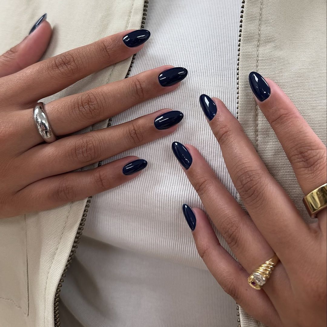 Primer plano de manos con manicura azul oscuro, cazadora beige y joyas doradas