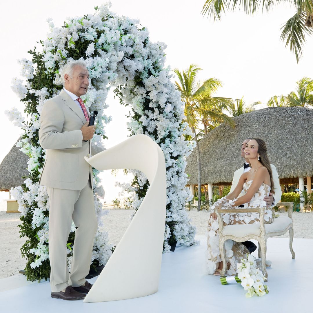 Hombre en traje claro y mujer en vestido floral sentados en playa con arco floral.