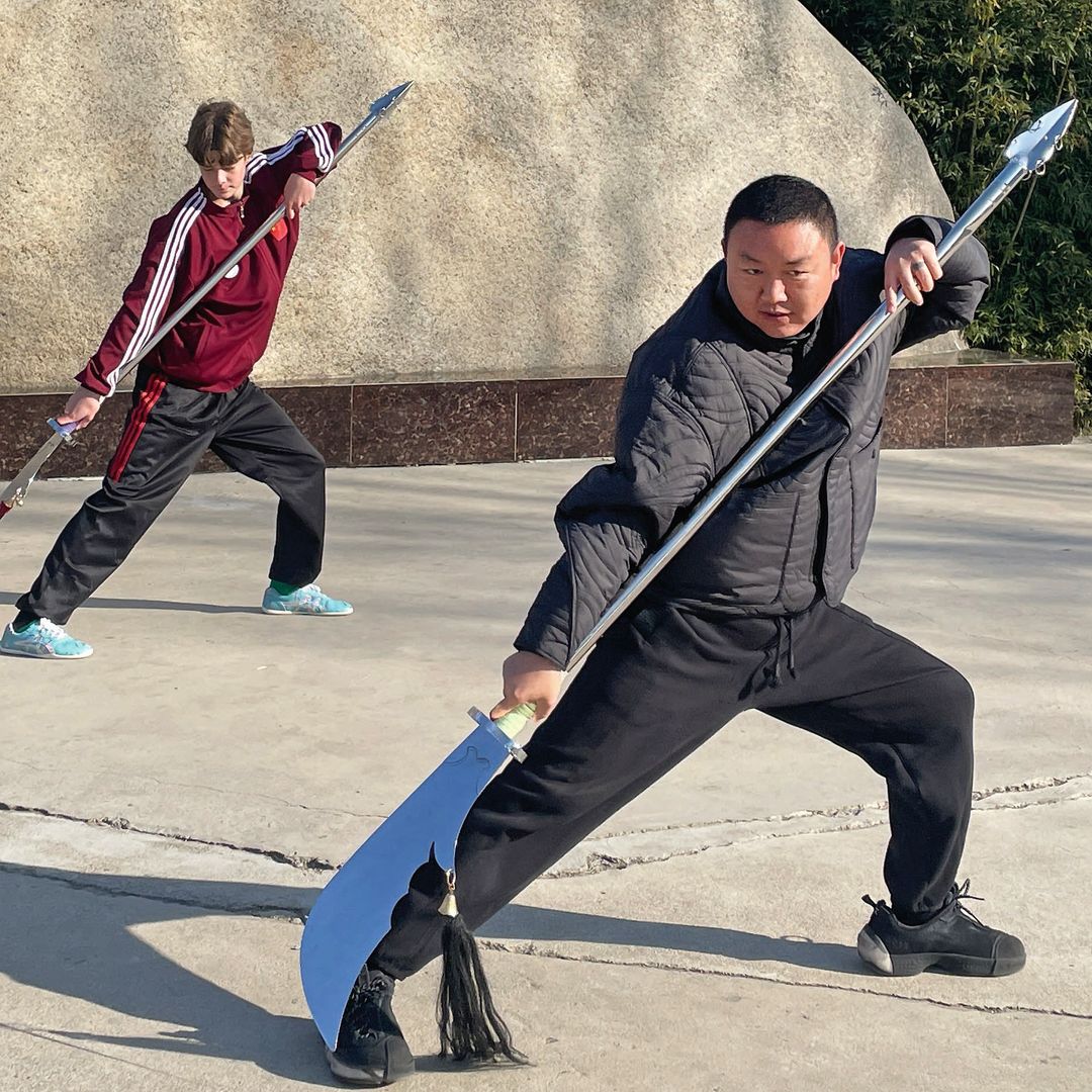 Simeón junto a otra persona practicando artes marciales con sables frente a una gran roca.