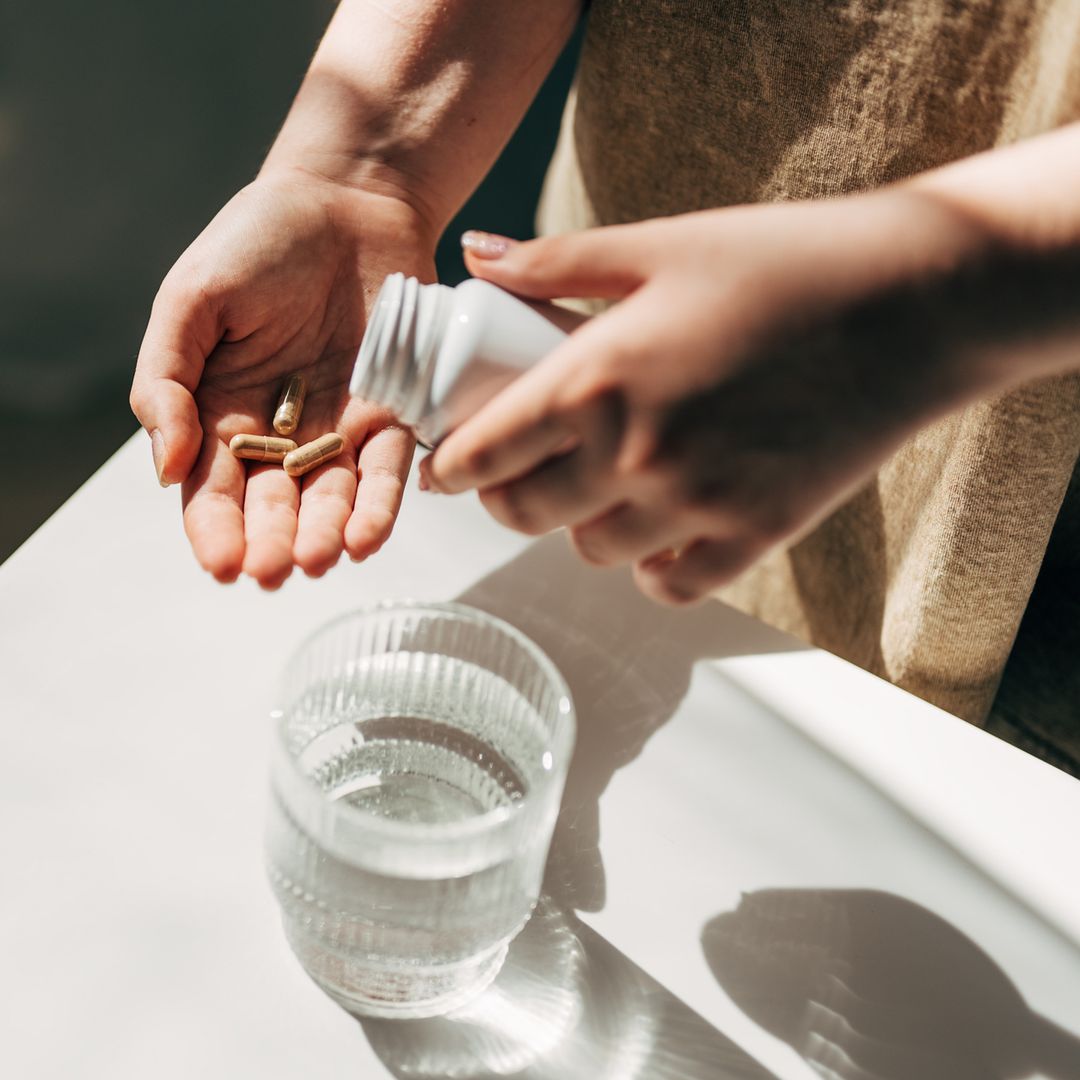 Mujer cogiendo unas pastillas de suplemento alimenticio con un baso de agua. Plano detalle de las manos.