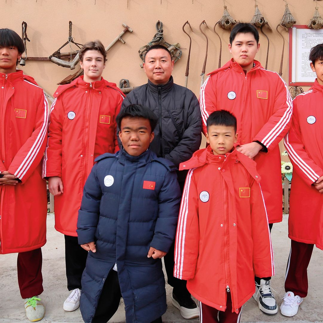 Simeón junto a ocho personas en trajes rojos y dos niños en abrigos azules posan frente a un edificio con escritura china.
