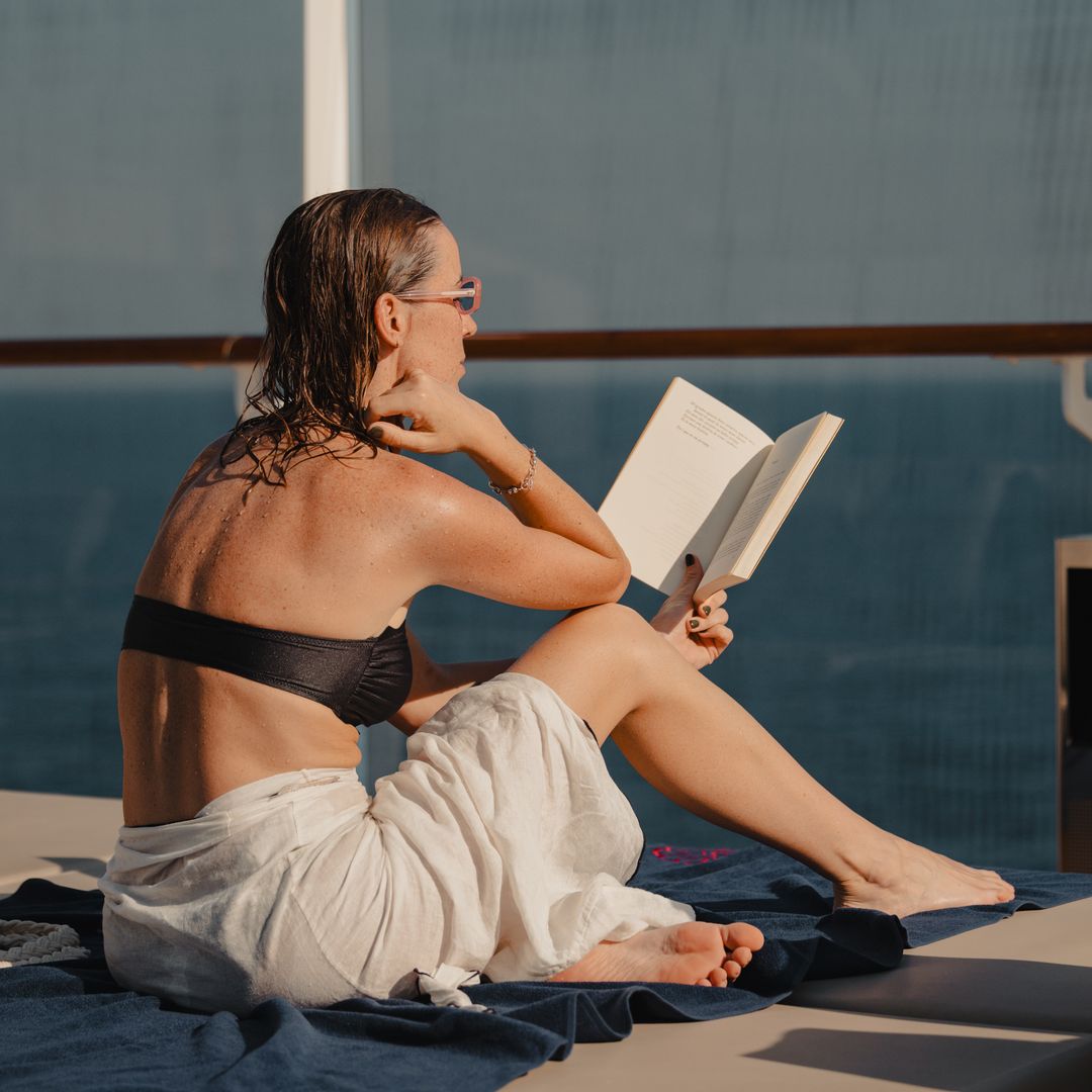 Mujer leyendo un libro en la cubierta de un crucero de Explora Journeys al atardecer, en bañador y con la toalla.