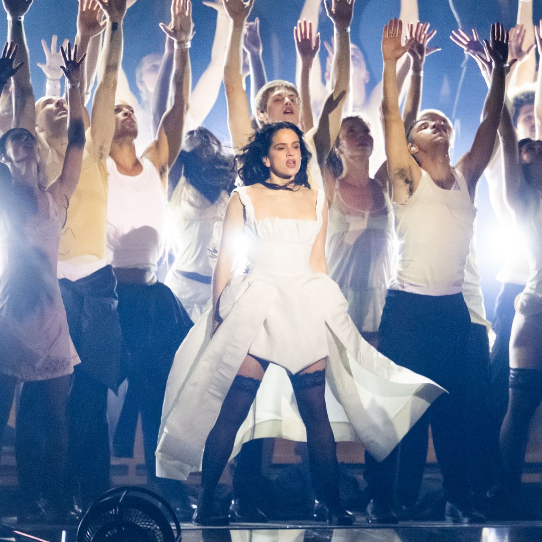 Rosalía, en un vestido blanco, actúa en un escenario con un coro elevando las manos.