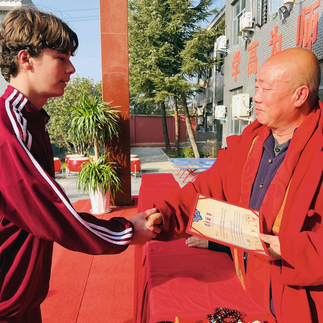 Simeón en sudadera roja estrechando la mano de un hombre mayor en túnica roja.