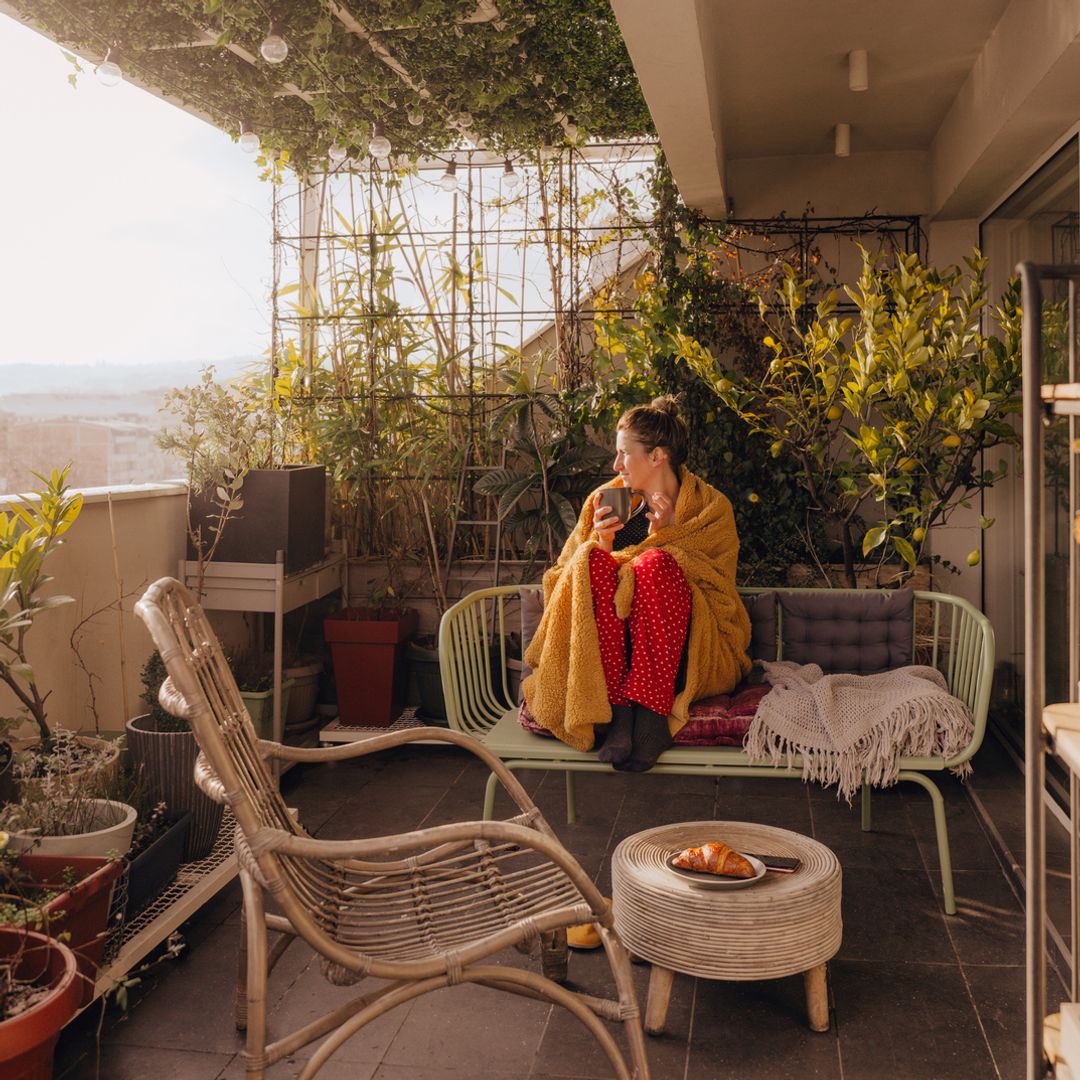 Chica tomándose una taza de café en su terraza con una manta sobre los hombros mientras mira el amanecer.