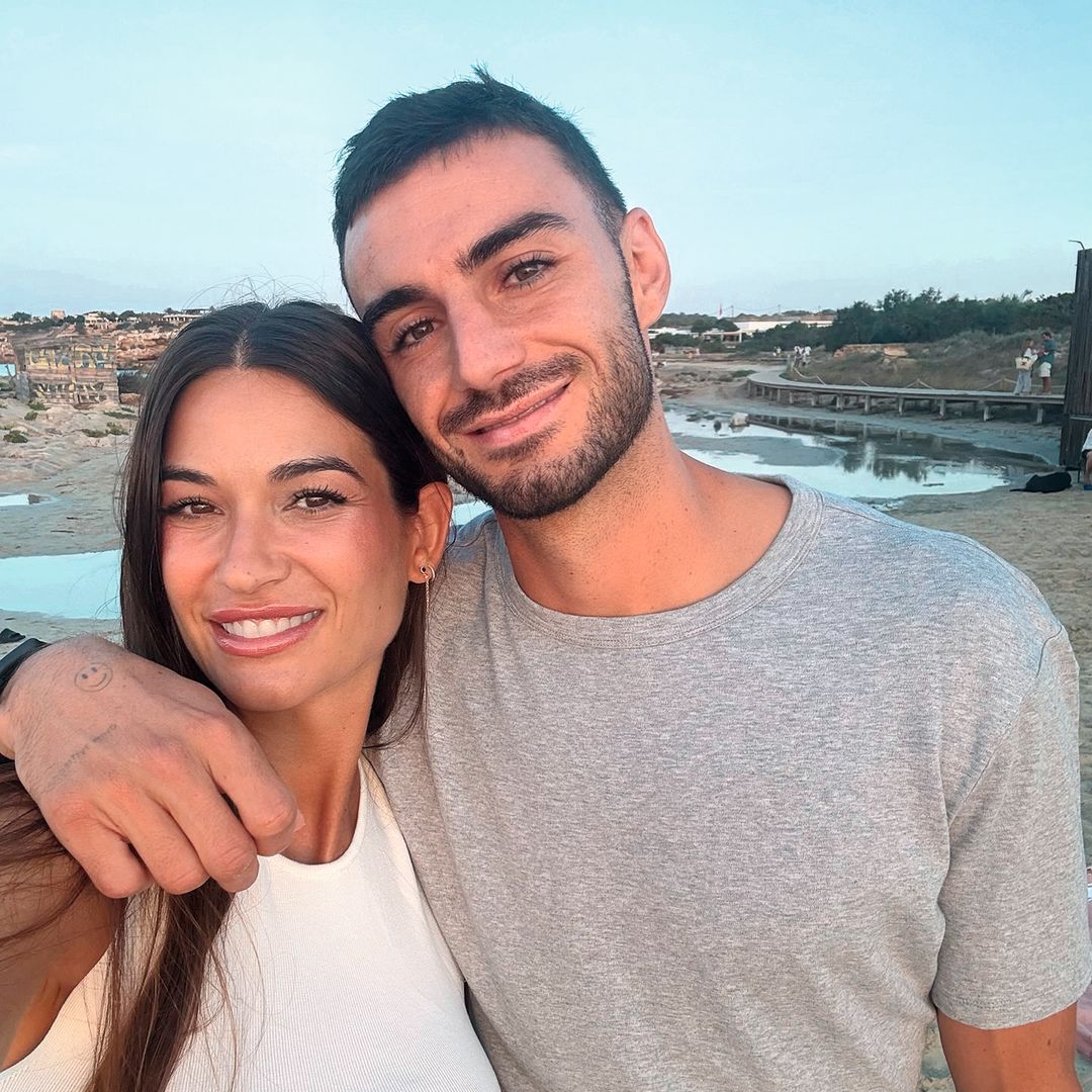 Estela Grande con vestido blanco.y Juan Iglesias con barba y camiseta gris en la playa, sonrientes