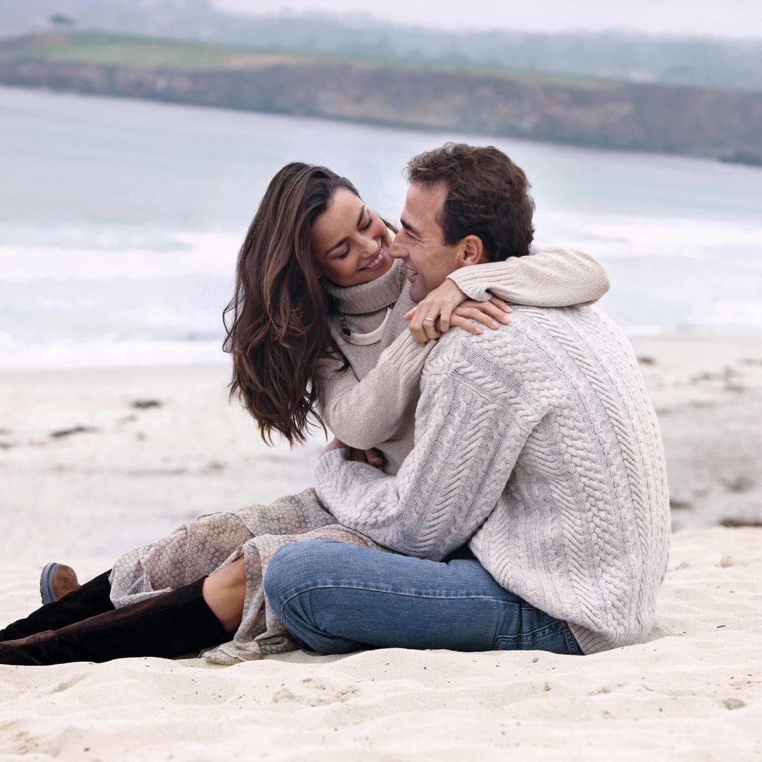 María Palacios y Alessandro Lequio abrazados sentados en la arena frente al mar, riendo y disfrutando del momento.