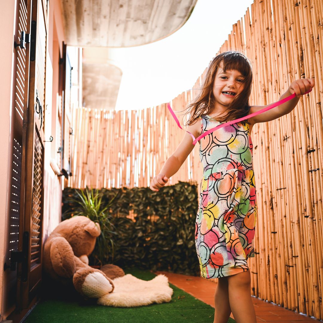Niña sonriente feliz en la terraza de casa jugando con una tira de color fucsia, con un oso de peluche.