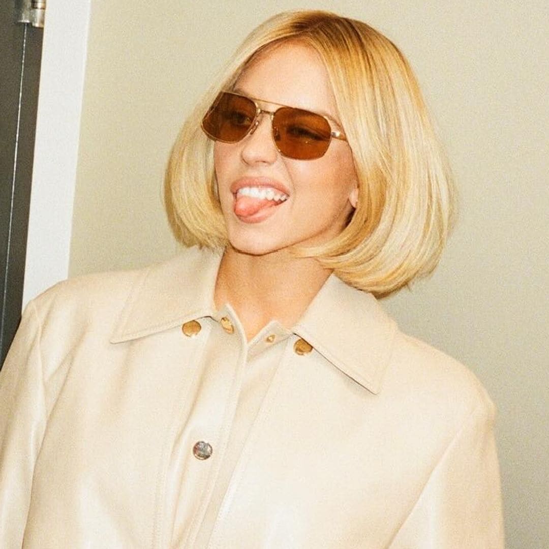 Sydney Sweeney con gabardina blanca de tejido brillante, gafas de sol marrones y corte de pelo a la altura de la mandíbula