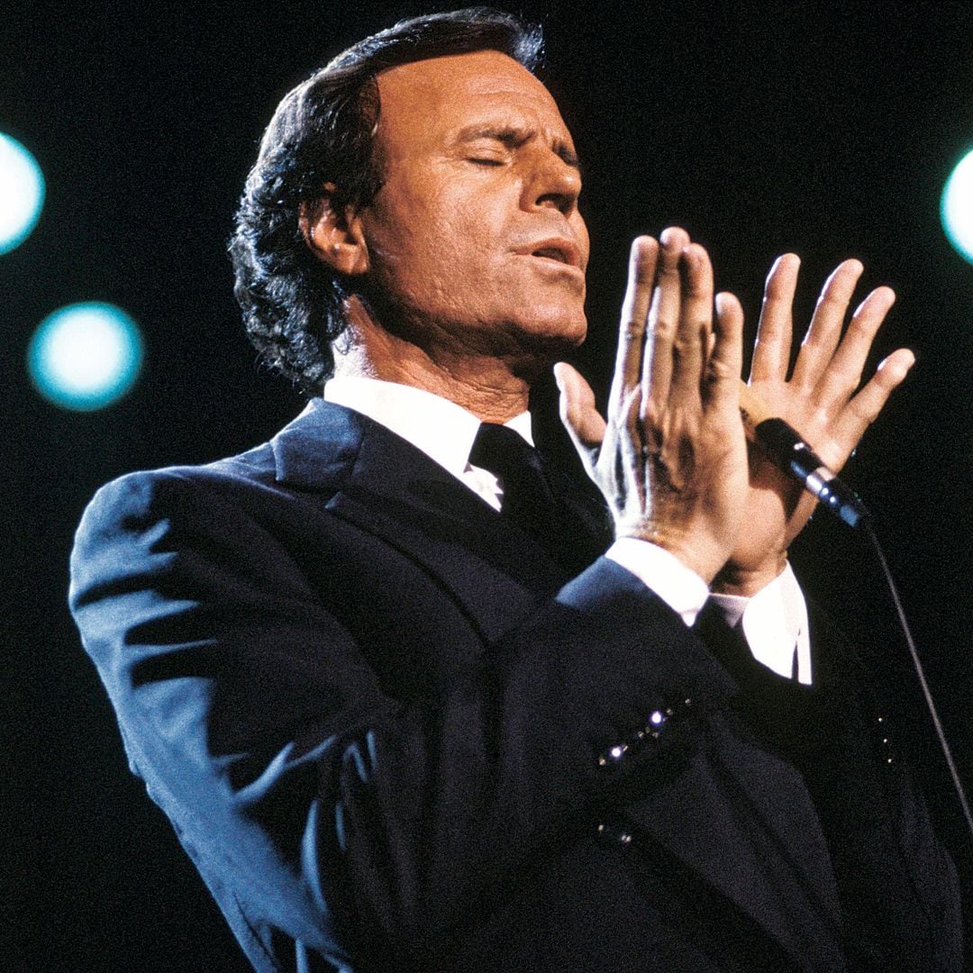 Julio Iglesias en un escenario, cantando con los ojos cerrados y las manos alzadas.