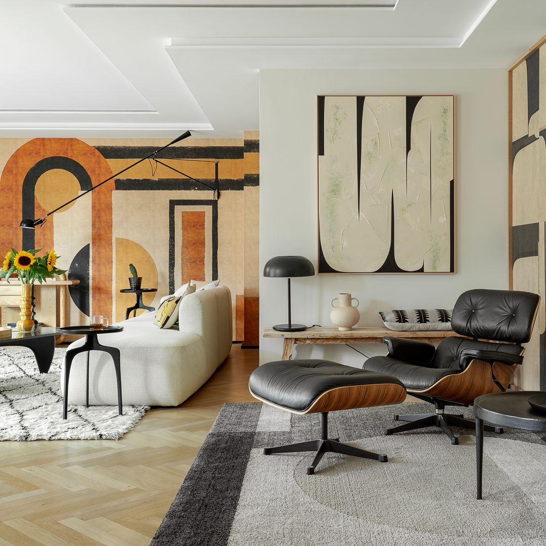 Salón con rincón de lectura, paredes con papel pintado con motivos geométricos y paneles, alfombras, chaise longue Eames, sofá blanco, mesa negra, jarrón con girasoles