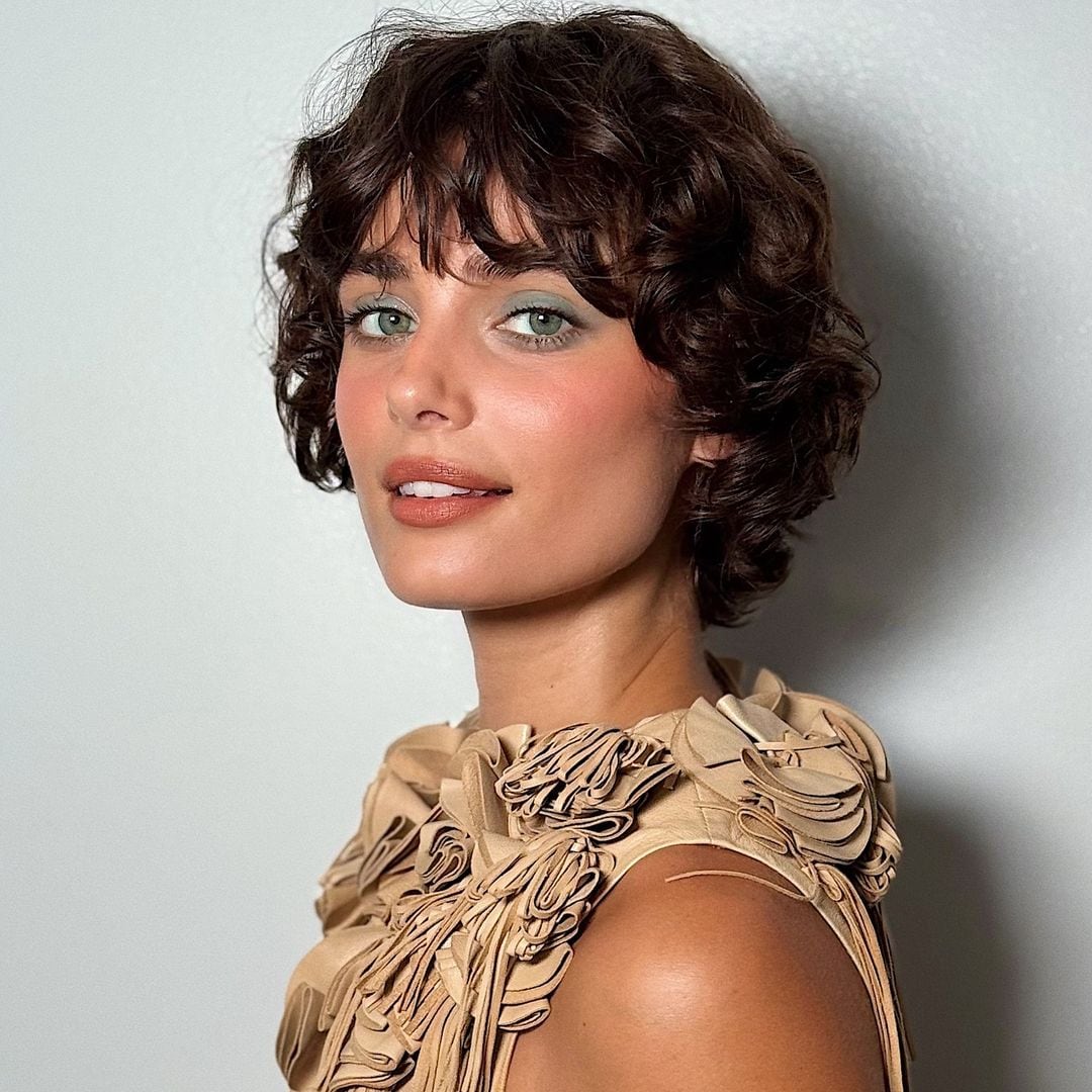 Taylor Hill con maquillaje verde, top de cuero beige y el pelo corto y rizado