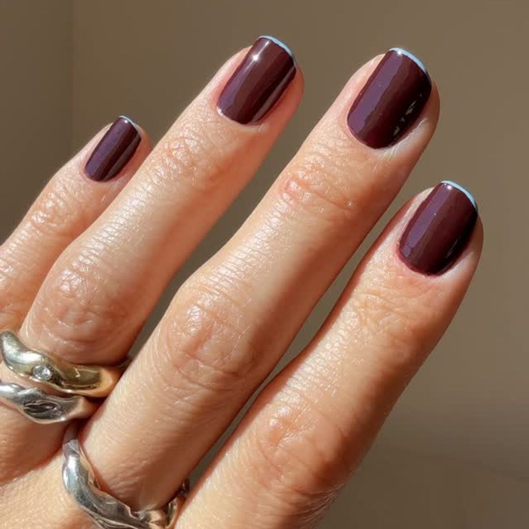 Mano con uñas pintadas en marrón oscuro y dos anillos plateados.