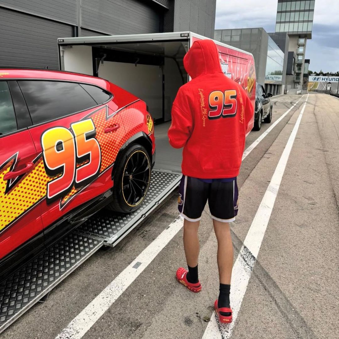 Plex con sudadera roja y capucha '95' junto a auto de carreras rojo en pista.
