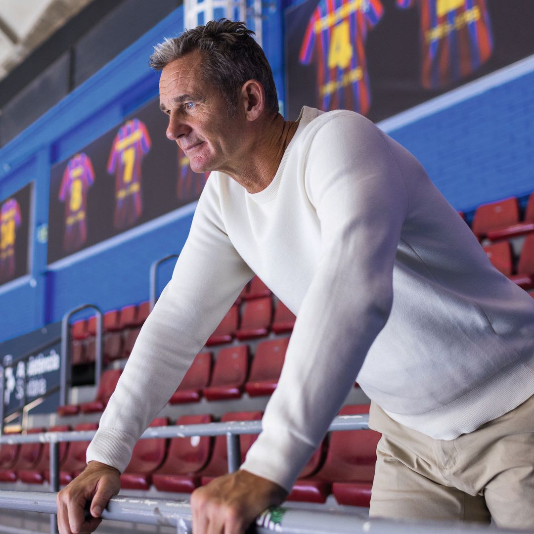 Iñaki Urdangarin en un estadio, apoyado en una barandilla, vistiendo una sudadera blanca y pantalones beige.