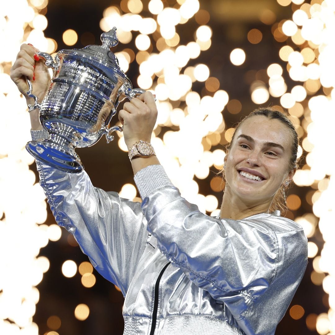 Aryna Sabalenka, con chaqueta plateada, sostiene un trofeo y sonríe con bengalas al fondo.