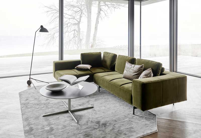 Salón con grandes ventanales con sofá verde oliva, alfombra gris y lámpara de pie