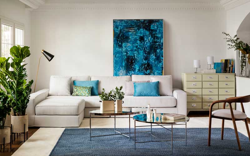 Salón con sofá blanco, plantas en el suelo y cojines y cuadro abstracto en color azul 