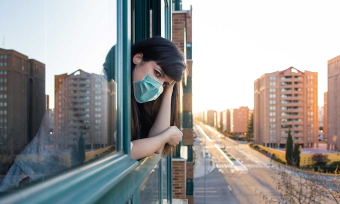 Una mujer con mascarilla asomada a la ventana con cara de preocupación por el confinamiento