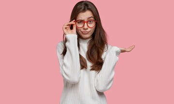 Chica joven con jersey blanco sobre fondo rosa. Con gafas mira con gesto divertido a cámara