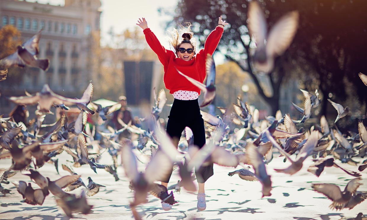 Mujer dando un salto de alegría en medio de una plaza rodeada de palomas