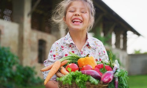 Una niña con cesta de verduras sonriendo en el campo. Cómo conseguir que los niños coman de manera saludable.